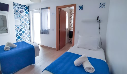 Quarto luminoso no Ó da Casa em Peniche com camas azuis, almofadas brancas, parede de destaque em azulejo português e porta com vista para o jardim