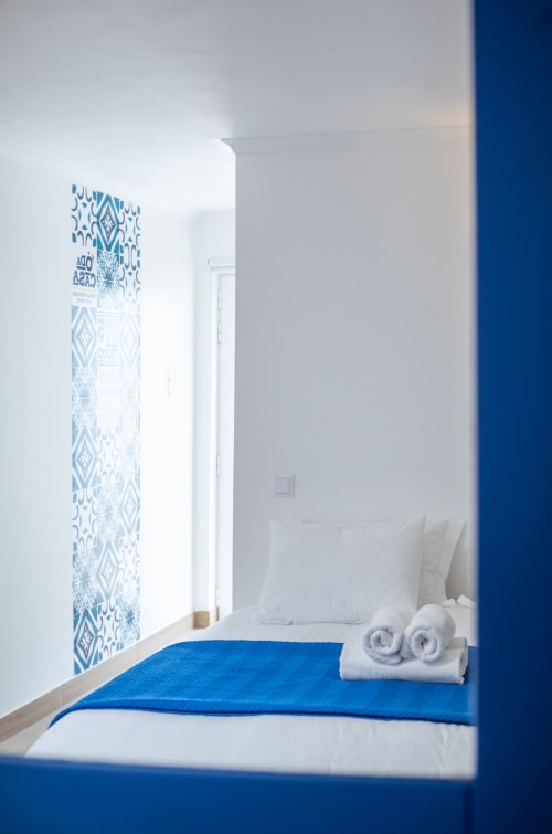 Quarto luminoso em Ó da Casa em Peniche com detalhes azuis, almofadas brancas, toalhas enroladas e arte de parede geométrica azul