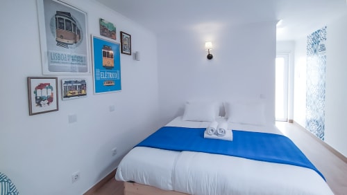 Quarto luminoso na hospedaria de Peniche com roupa de cama azul e branca, cartazes de elétrico português e vistas para o jardim