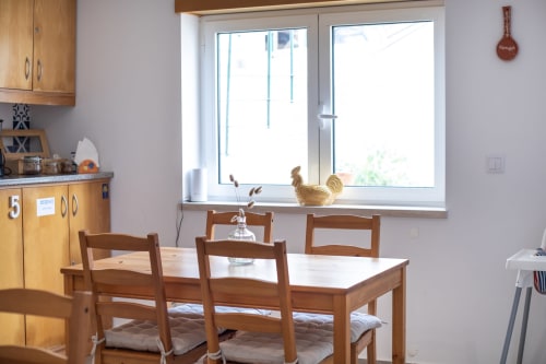 Cuisine-salle à manger lumineuse à Ó da Casa B&B à Peniche avec table en bois, chaises et figurine de coq sur le rebord de fenêtre
