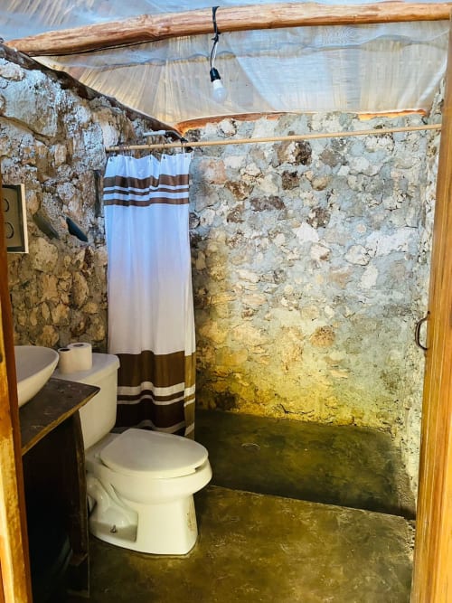 Baño rústico en el hostal Cayuco Maya de Bacalar con paredes de piedra, inodoro, lavamanos y cortina de ducha a rayas