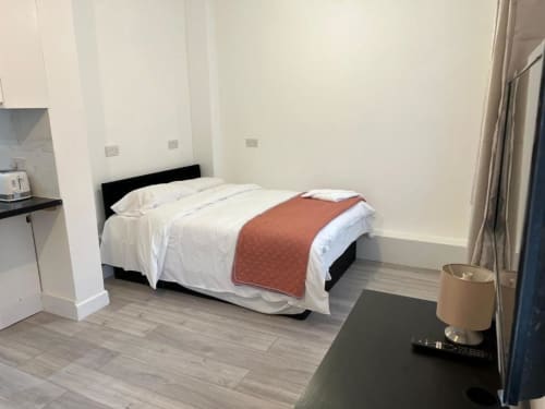 Modern hotel room at Starlet Property London with queen bed, white linens, orange throw, kitchenette, and neutral decor