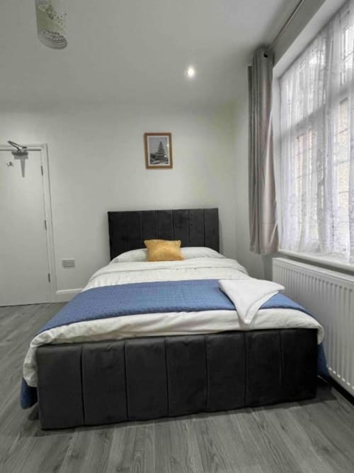 Modern en-suite bedroom at Starlet Property London with black bed, white linens, large windows, and wooden flooring.