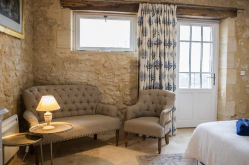 Chambre élégante du Château Haut Clarise avec murs en pierre, canapé capitonné, fauteuil et portes-fenêtres à Belvès-de-Castillon