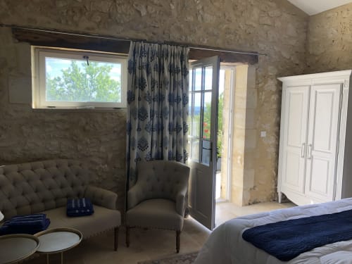 Chambre élégante au Château Haut Clarise avec murs en pierre, rideaux bleu à motifs, sièges rembourrés et portes sur jardin