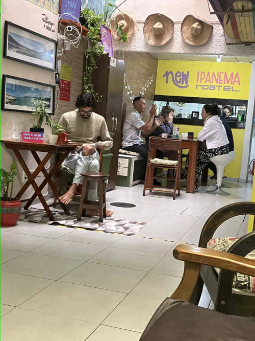 Área comum do Hostel New Ipanema com hóspedes sentados em mesas de madeira, Rio de Janeiro