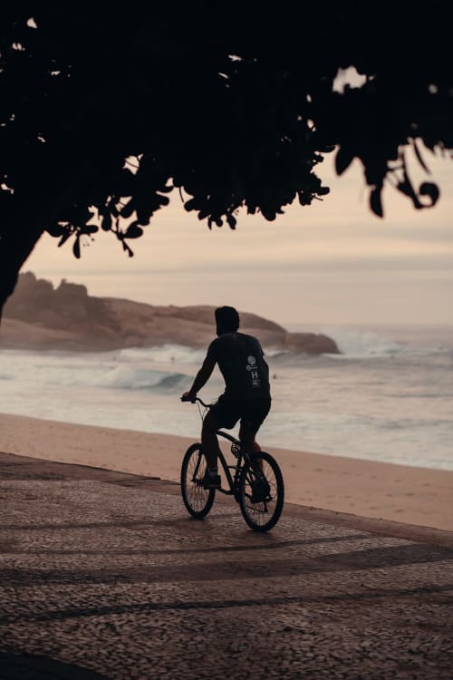 Ciclista pedalando na praia do Rio ao pôr do sol com ondas e vegetação tropical acima