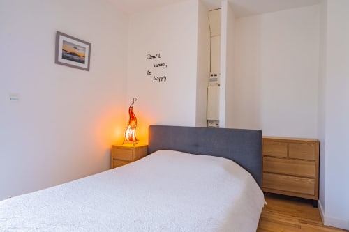 Chambre douillette d'un appartement à Toulouse avec lit blanc, table de nuit en bois, lampe rouge lumineuse et texte de motivation au mur
