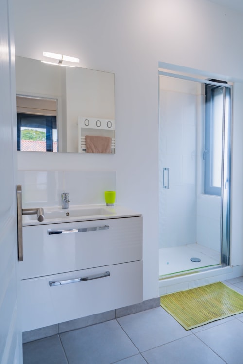 Salle de bains moderne dans un appartement toulousain avec vasque blanche, miroir, douche vitrée et tapis de bain vert