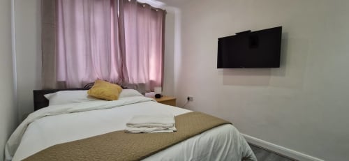 Starlet Property London hotel bedroom with queen bed, white linens, yellow pillow, wall-mounted TV, and pink curtains