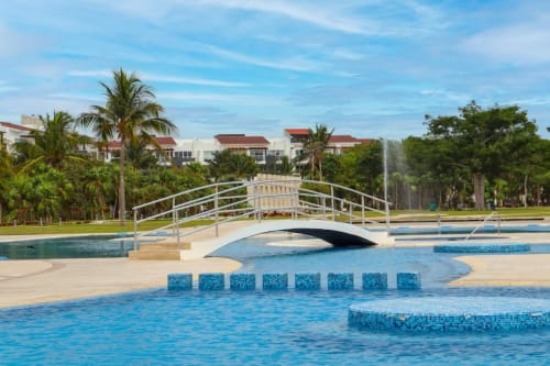 Lujosa zona de piscina del Mareazul Beachfront Resort con puente blanco, azulejos azules y palmeras en Playa del Carmen