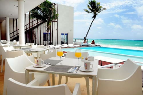 Área de comedor frente al mar de Mareazul Resort con sillas blancas, vista al océano turquesa, palmeras y arquitectura moderna en Playa
