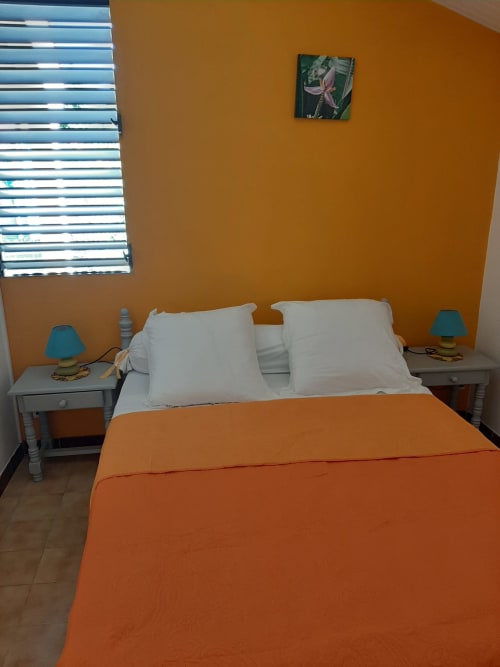 Bright bedroom at Mon Refuge in Martinique with orange walls, white pillows, orange bedcover, turquoise lamps, and louvered