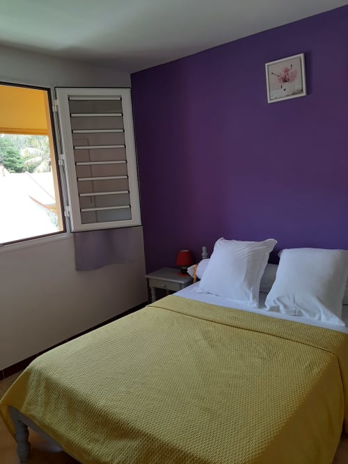 Chambre lumineuse aux murs violets, couvre-lit jaune, oreillers blancs et vue sur le jardin à Les Anses d'Arlet