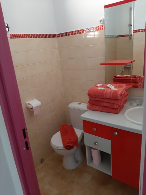 Salle de bain moderne avec vanité rouge et blanc, carrelage crème, serviettes rouges et toilettes à l'appartement Mon Refuge aux Anses