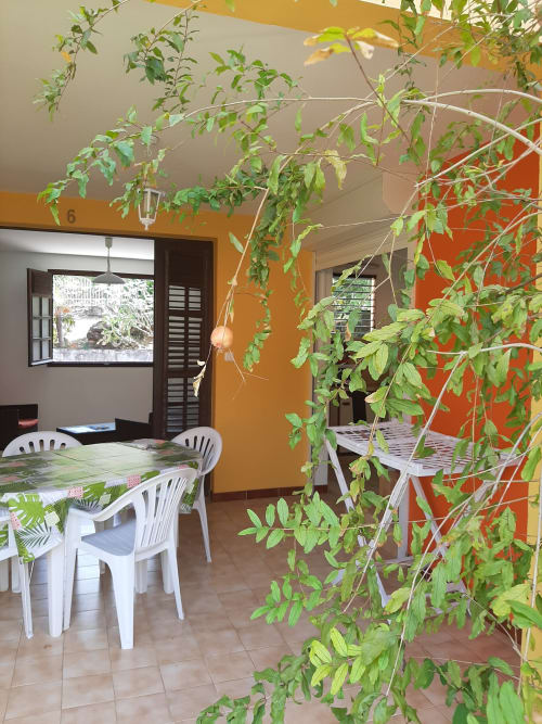 Coin repas à la Résidence de Vacances Mon Refuge avec chaises blanches, plantes suspendues luxuriantes et murs aux teintes jaune et orange éclatantes
