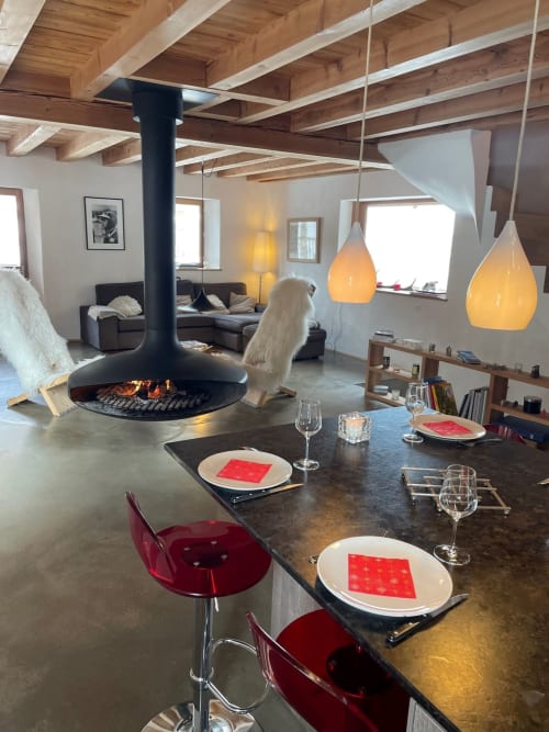 Intérieur de chalet moderne à Tignes avec foyer suspendu, table à manger, chaises rouges et luminaires ambre sous poutre apparente