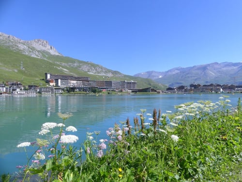 Lac alpin turquoise avec fleurs sauvages blanches et roses, montagnes et bâtiments de la station de Tignes sous ciel bleu dégagé