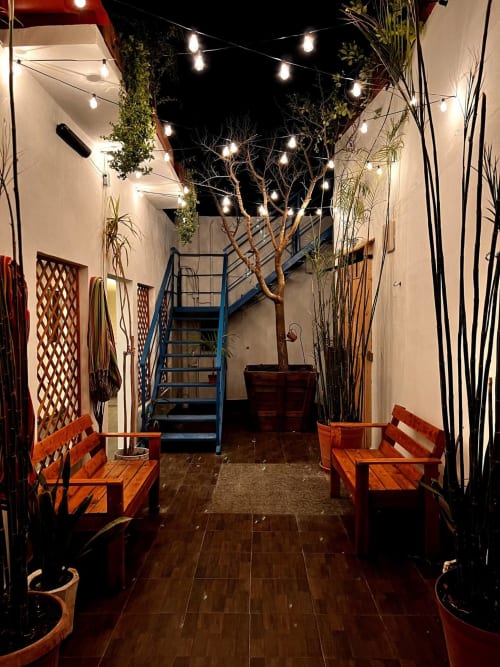 Patio del Hotel Boutique BajaReal por la noche con luces colgantes, bancos de madera, plantas en macetas y escalera metálica azul en La