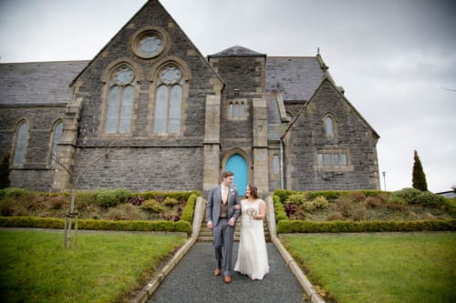 Bride and groom standing on pathway in front of historic stone church at Sliabh Beagh Hotel in Monaghan, Ireland