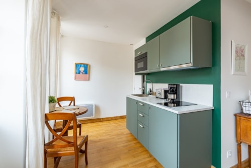 Modern kitchenette with sage green cabinetry, white countertops, and wooden dining area in Logis Jeanne apartment at La