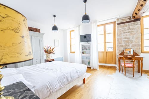 Bright bedroom at La Maison George with white bed, yellow lamp, wooden desk, stone walls, and modern black pendant lights