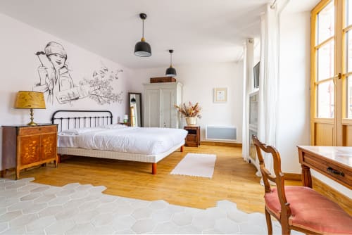 Spacious bedroom at La Maison George featuring a metal bed frame, wooden furniture, sketch wall art, and large windows with