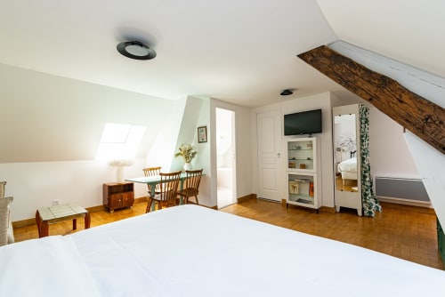 Chambre mansardée spacieuse à La Maison George avec lit blanc, poutres apparentes, télévision, coin repas et lucarne