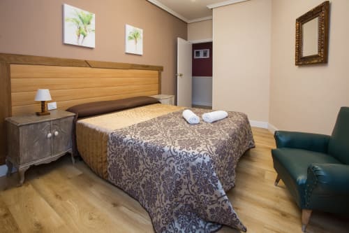 Bright bedroom at Apartamentos Turísticos Ávila with wooden bed, nightstand lamp, green armchair, and hardwood flooring