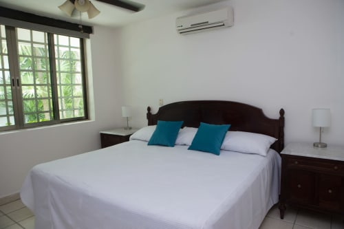 Acogedora habitación en casa del Centro de Cancún con cama de madera oscura, almohadas turquesas, ropa blanca, aire acondicionado y vistas al jardín.