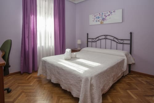 Dormitorio luminoso en Apartamentos Turísticos Ávila con cortinas moradas, colcha blanca, cama de hierro ornamentada y piso de madera