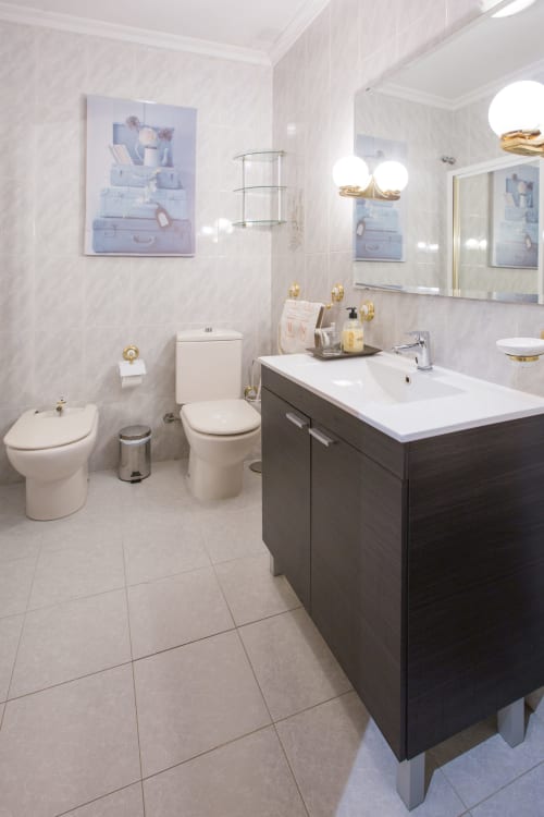 Baño moderno en apartamento vacacional de Ávila con azulejos blancos, tocador oscuro, doble lavabo y accesorios dorados