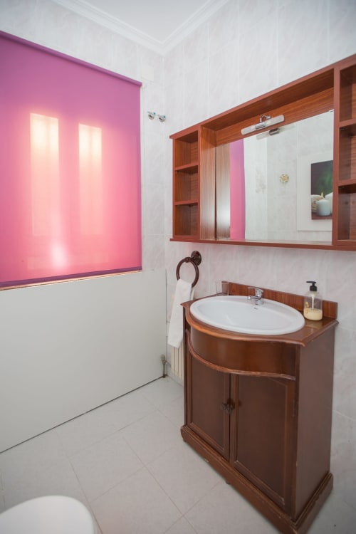 Baño moderno con mueble lavabo de madera, piso de azulejos blancos, pared de acento rosa y estantes con espejo en Apartamentos Turísticos