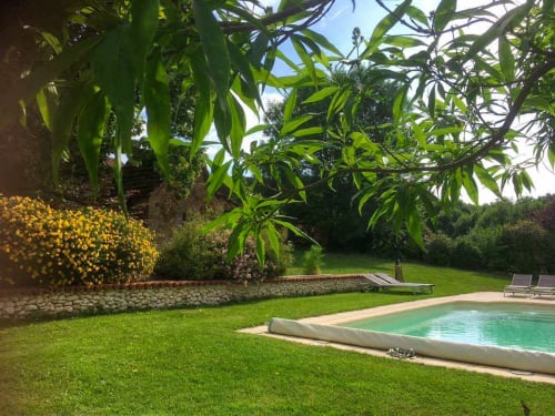 Jardin ensoleillé avec piscine, pelouse verte, plantes fleuries et mur en pierre rustique au gîte Le Lapin sur le Toit
