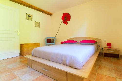 Chambre lumineuse au Gîte La Grange avec lit en bois, oreillers roses, coquelicot rouge et carrelage en terre cuite