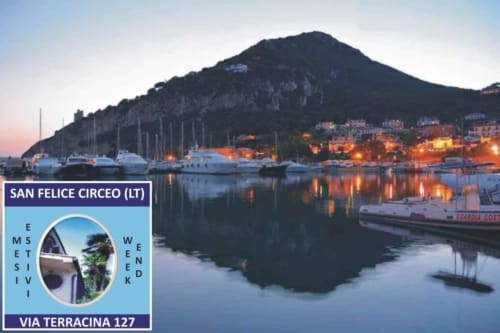 Porto di San Felice Circeo al tramonto con edifici illuminati, barche a vela e montagne sullo sfondo nel Lazio, Italia