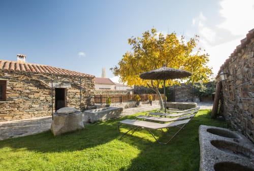 Patio rústico de piedra en casa rural de Ávila con tumbonas, sombrilla y árbol dorado de otoño de fondo