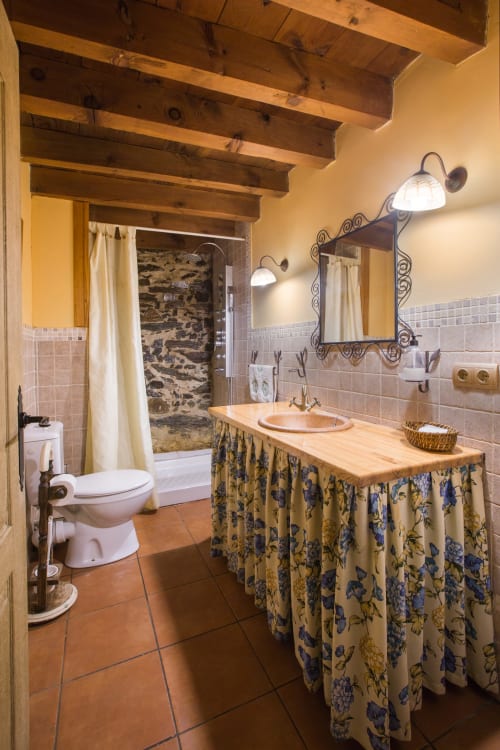 Baño rústico en casa rural El Horno de Rinconada, Ávila, con vigas de madera, baldosas de barro y lavabo decorativo con motivos florales