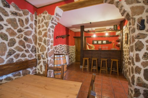 Zona de bar rústica con paredes de piedra y blancas, acentos rojos, vigas de madera, taburetes y decoración tradicional en Apartamentos