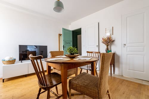Logis Colette dining room with wooden table, wicker chairs, TV, and green bathroom door at La Maison George