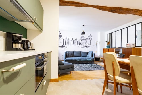 Modern open-plan apartment at La Maison George with green kitchen, blue sofa, dining area, and exposed wooden beams