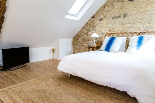 Chambre mansardée moderne avec mur en pierre, parquet, lit blanc aux coussins bleus, velux et télévision aux Filles du Bord de