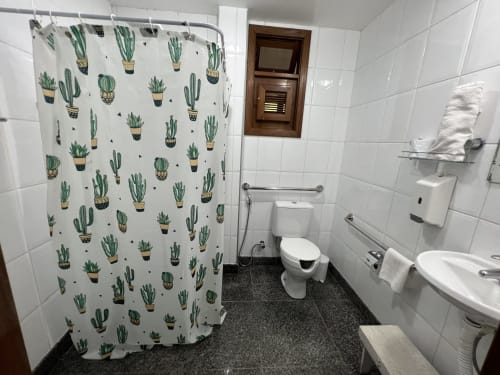 Banheiro limpo no Pouso do Barão com cortina de chuveiro estampada de cactos, vaso sanitário, pia e paredes azulejadas brancas em Domingos