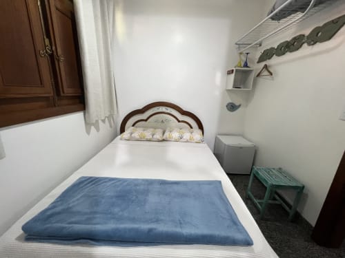 Quarto aconchegante no Pouso do Barão em Domingos Martins com cabeceira de madeira, roupa de cama branca, manta azul e janela com vista para o jardim