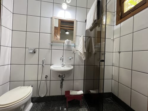 Banheiro moderno do Pouso do Barão com paredes de azulejo branco, pia, vaso sanitário, chuveiro e bancada escura em Domingos Martins