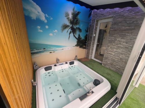 Bain à remous extérieur avec fresque tropicale, façade en brique et gazon synthétique du jardin du cottage Domérat