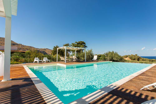 Turquoise swimming pool with wooden deck, white pergolas, and sea view at Villa Maria, Sant'Andrea Valderice Sicily