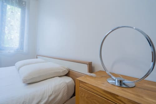 Chambre lumineuse dans un appartement toulousain avec literie blanche, table de nuit en bois et miroir rond reflétant la lumière de la fenêtre