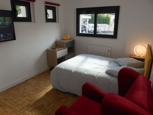Chambre moderne dans un appartement toulousain avec lit blanc, fauteuil rouge, mobilier en bois et vue sur la ville