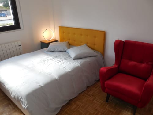 Chambre moderne dans un appartement toulousain avec tête de lit jaune, literie blanche, fauteuil rouge et parquet